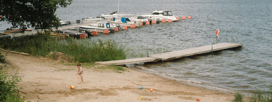 Badplats vid Mälaren. Badleksaker och ett litet barn på stranden. Brygga och båter i bakgrunden