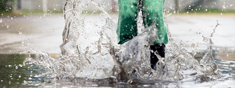 Ben med regnkläder och gummistövlar skvätter vatten i vattenpöl